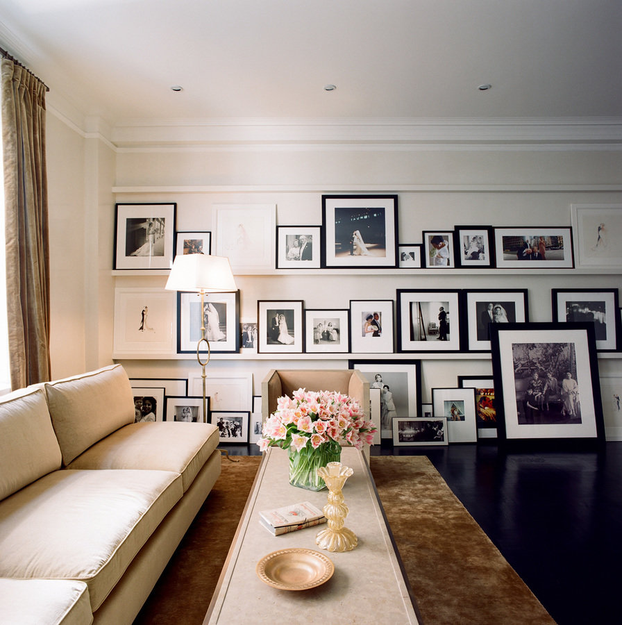 Living room with beige sofa at left, long coffee table with objects and flowers; back wall of shelves with framed photographs.