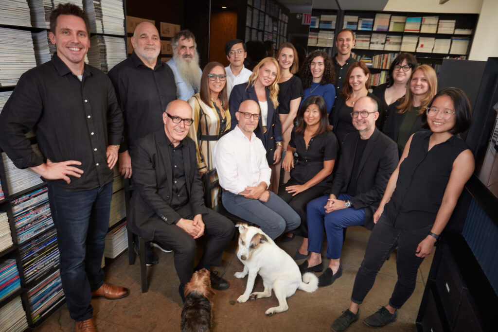 A group photo of men and women inside of an office with bookshelves in the background and a white dog in the foreground.