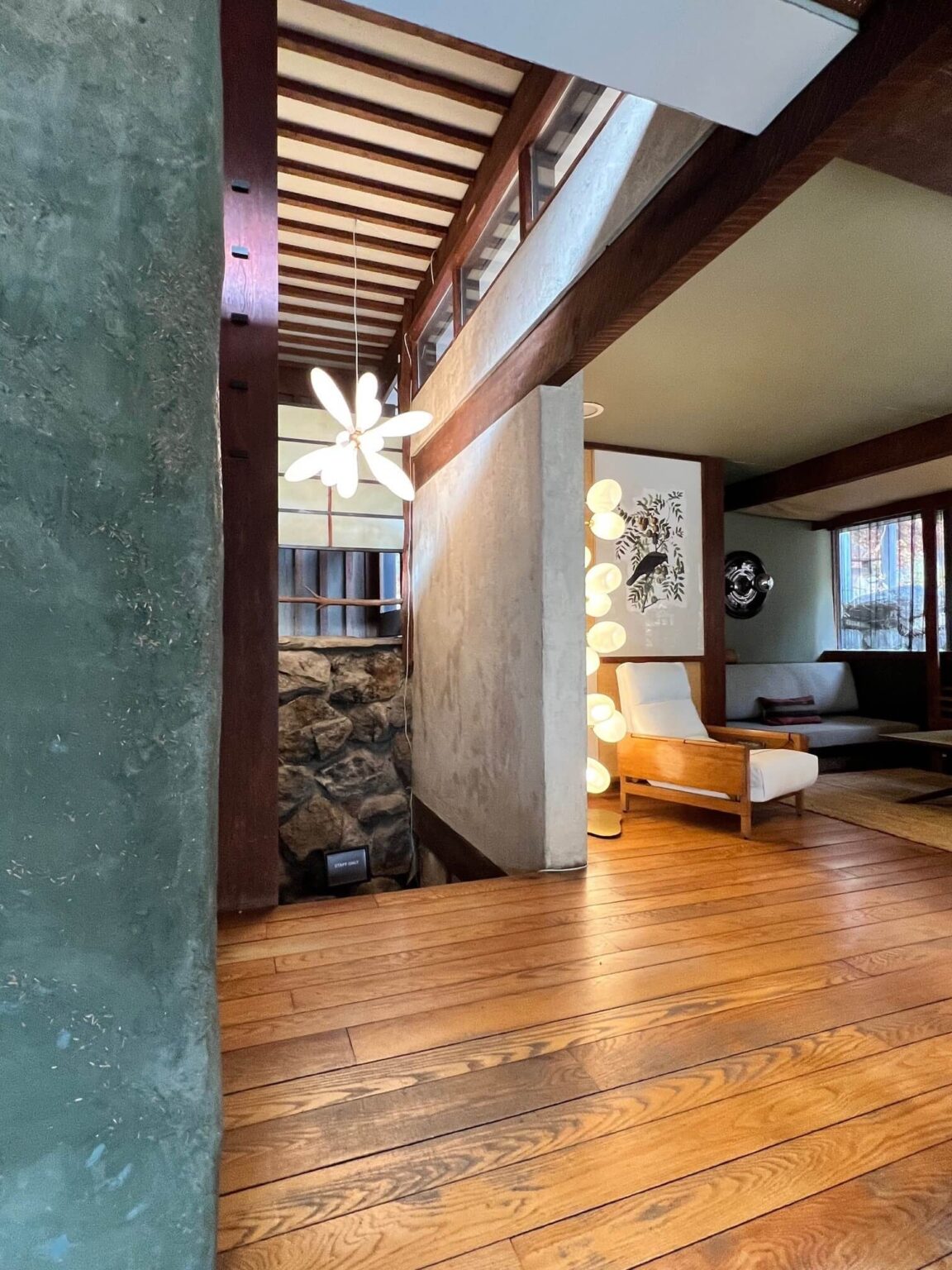 An interior with wood floors, a wood beam ceiling, organic light fixtures and a gray-green wall in the foreground.