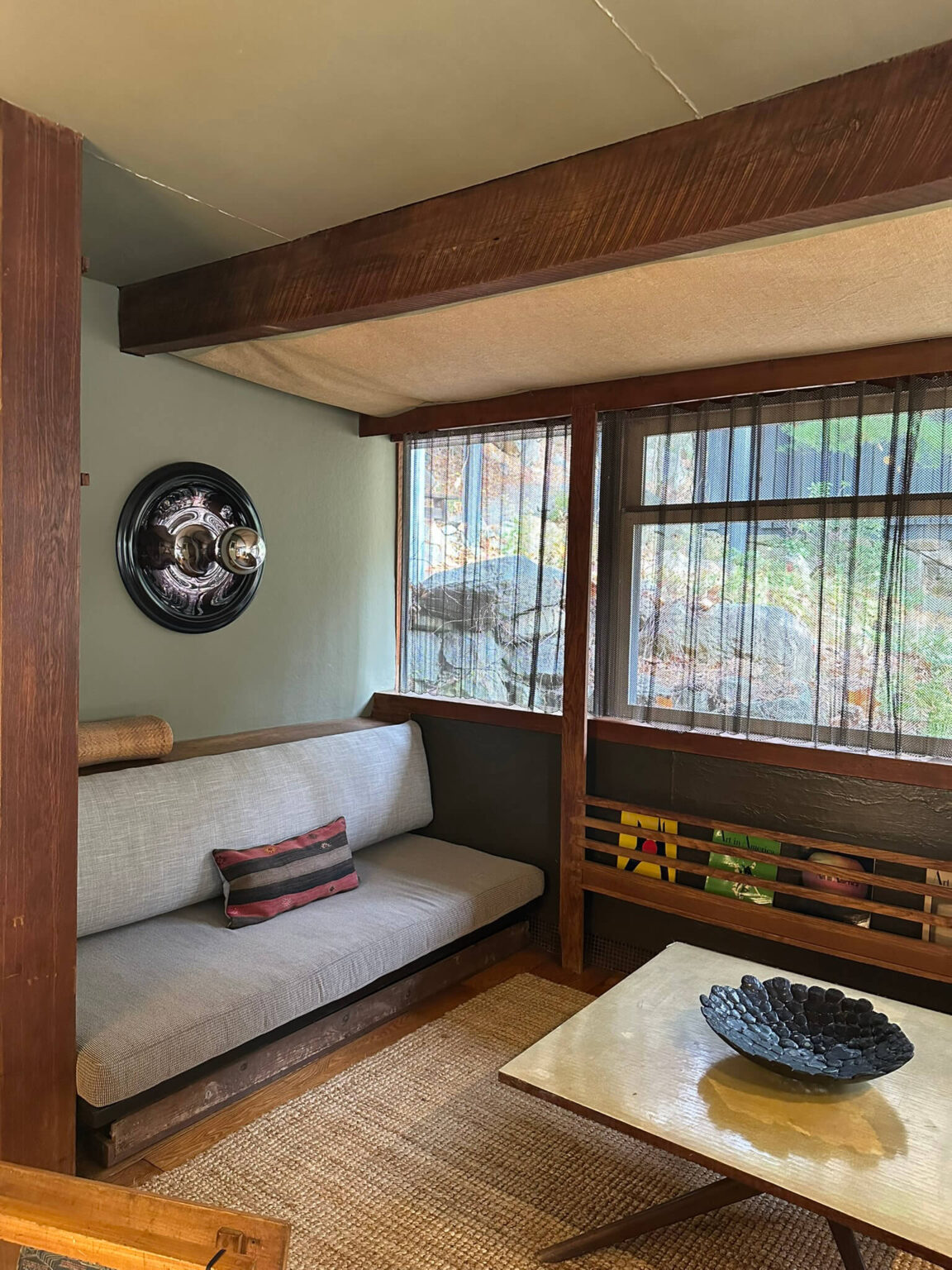 A den with a banquette sofa and square coffee table, an abstract, wall light, and windows with a view of rocky terrain.