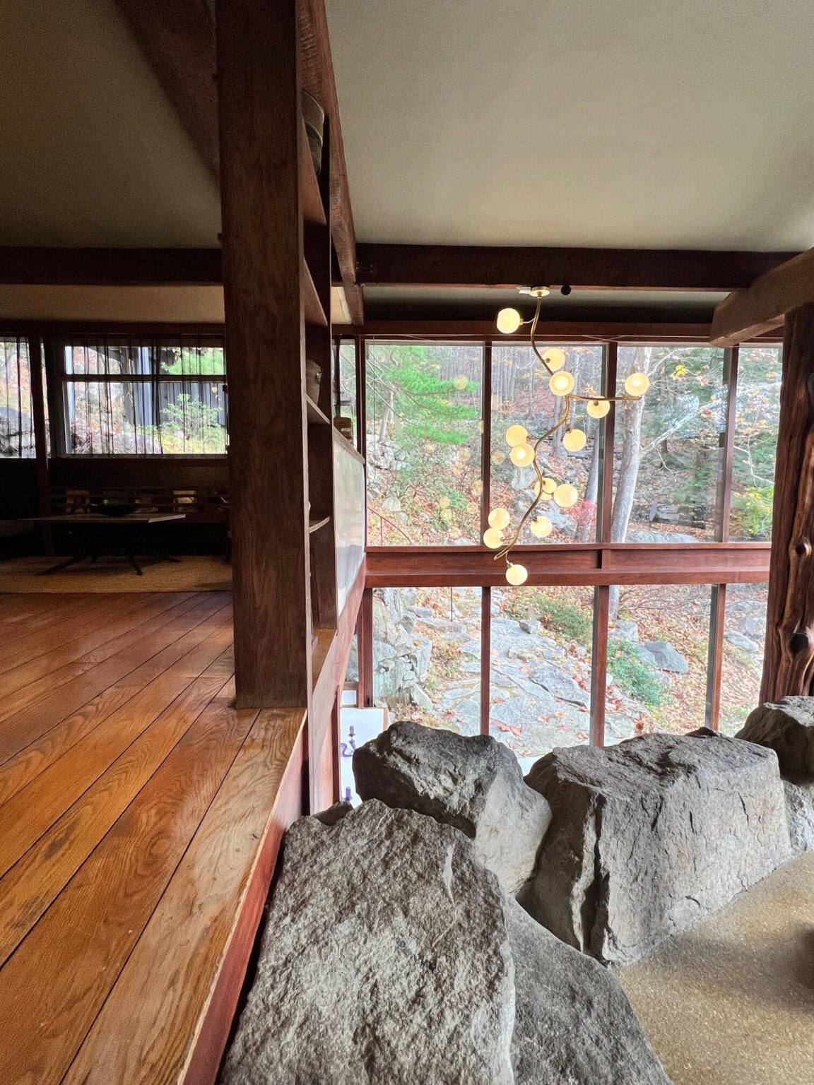 An interior view showing upper and lower levels of a house with a wall of windows, wood surfaces and a wall of boulders.