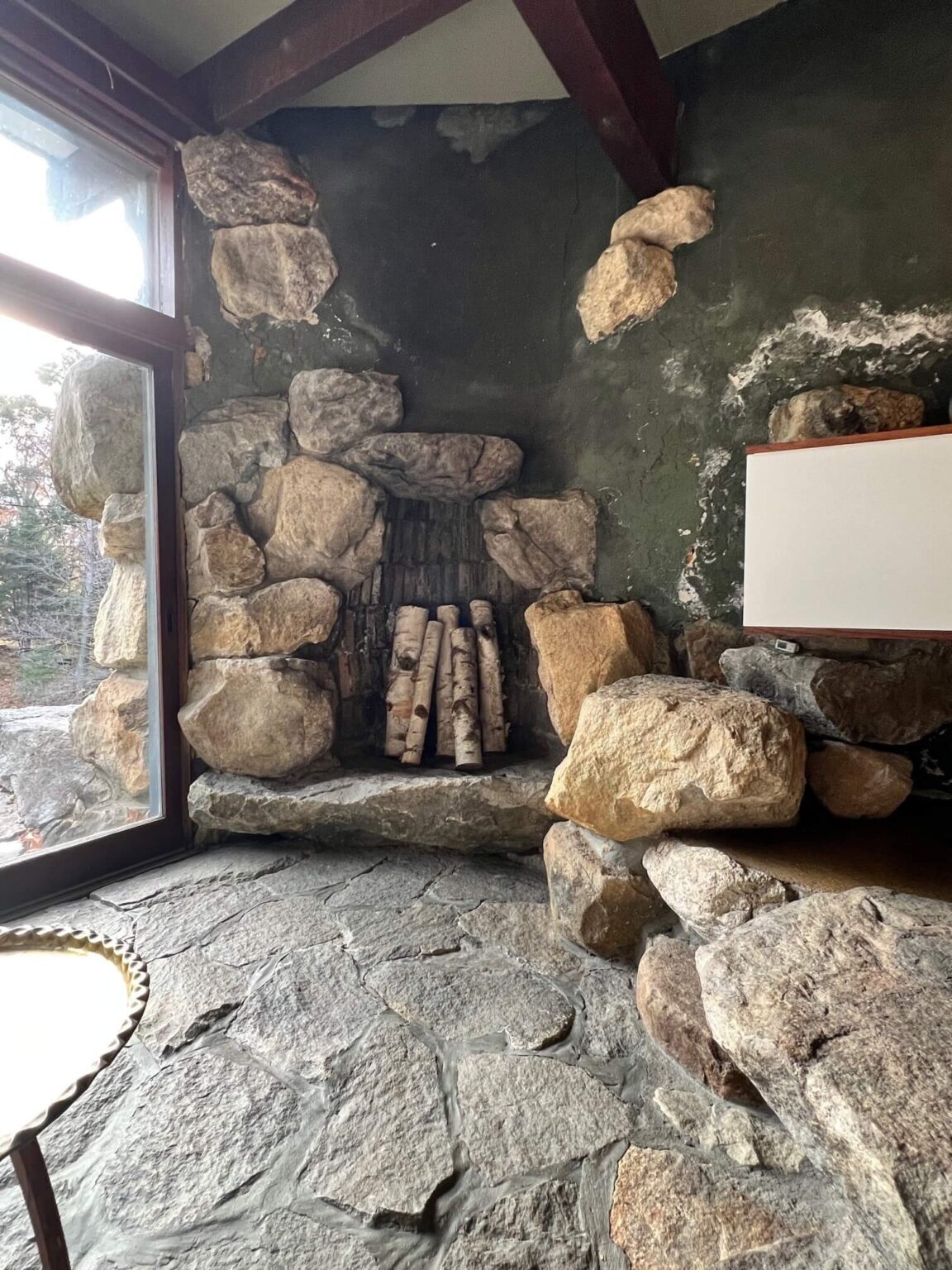 An interior with stone floors, and stacked boulders creating a fireplace, dark gray-green walls and windows at the left.