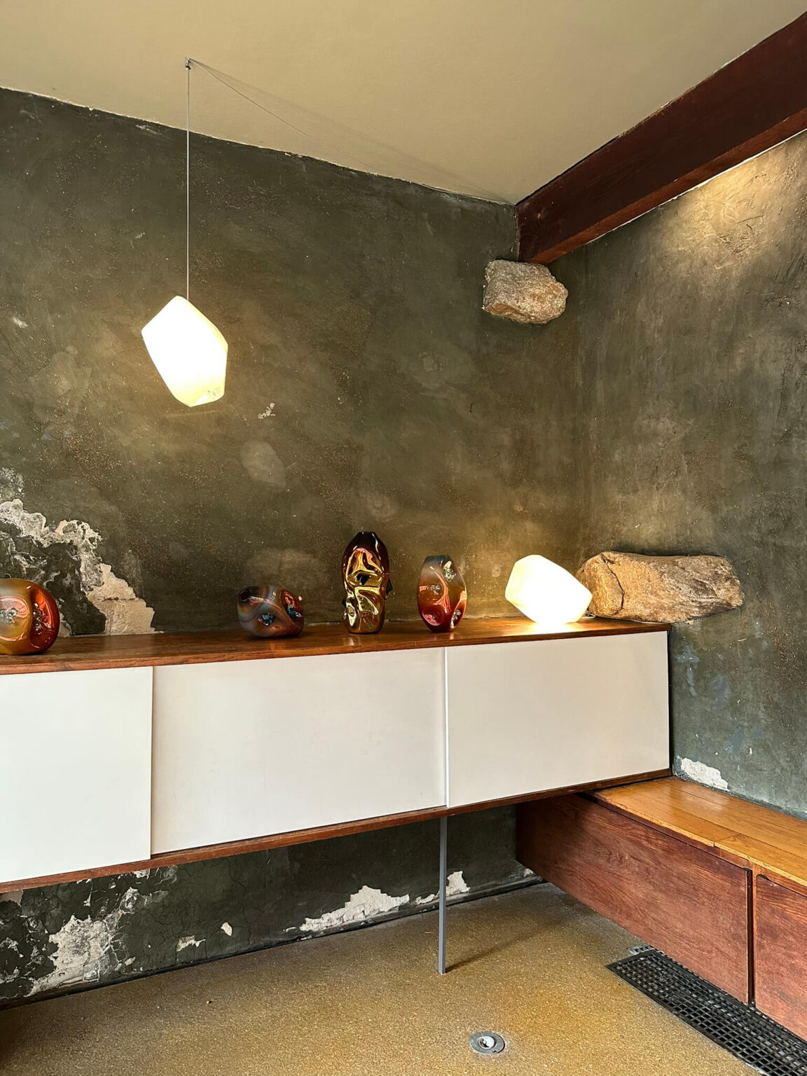 An interior with rough gray-green walls, a white and wood cabinet with shiny tabletop sculptures and a hanging light.
