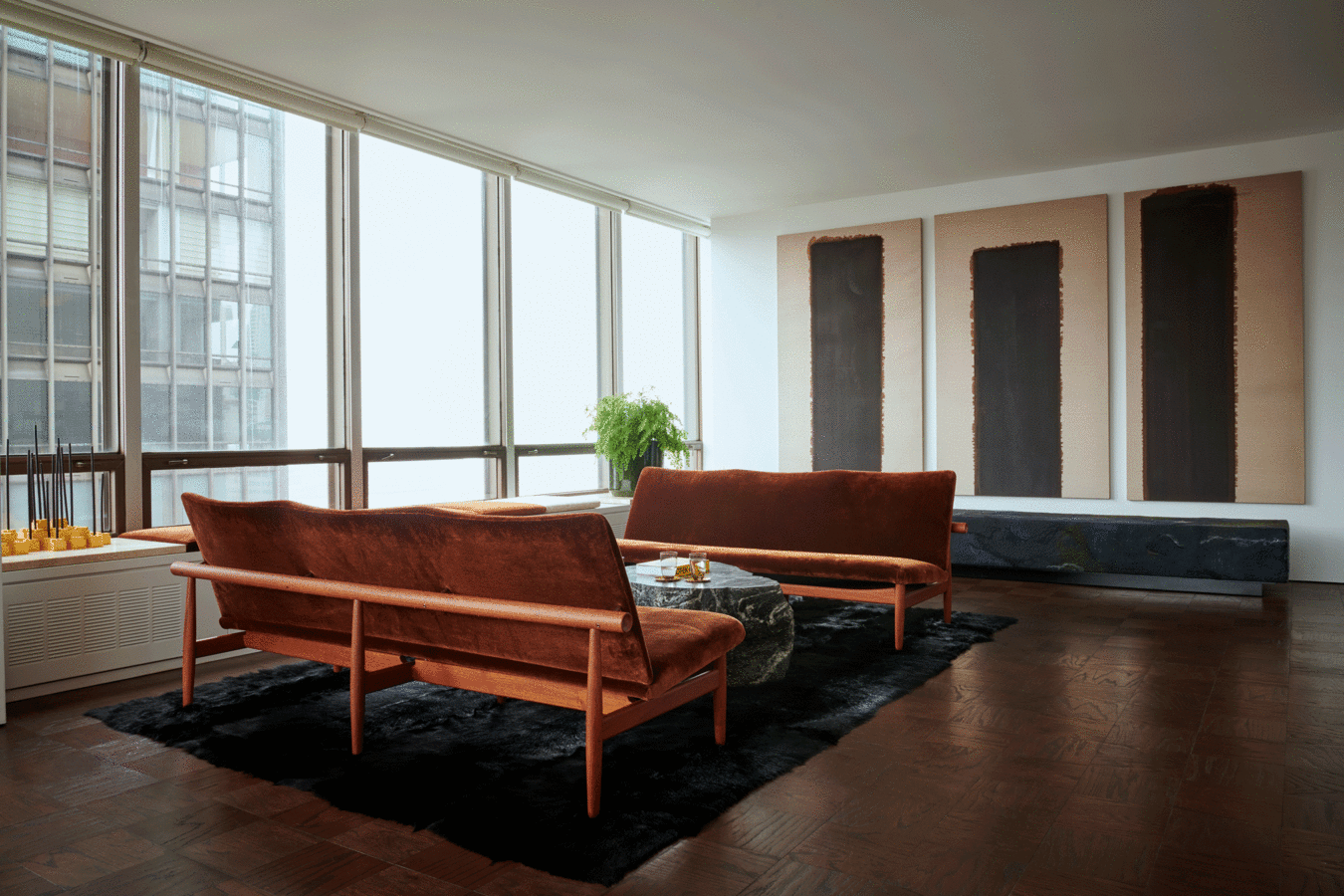 Living room with window wall, black and white triptych, wood floor, black rug and pair of wood settees with brown cushions.