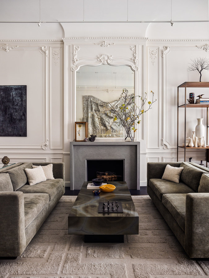 Prewar living room with fireplace and mirror, geometric pattern rug, pair of pale green velvet sofas and stone coffee table.