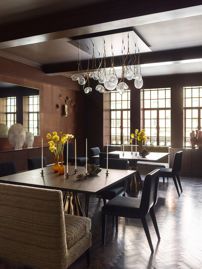 Dining room with brown leather walls, two tables, chairs, abstract glass chandelier, casement windows and chevron wood floor.