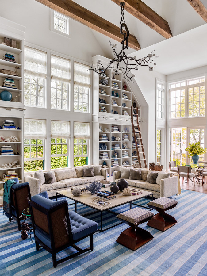 Double-height living room with white divided light windows, millwork shelving, exposed beams, seating area, blue striped rug.