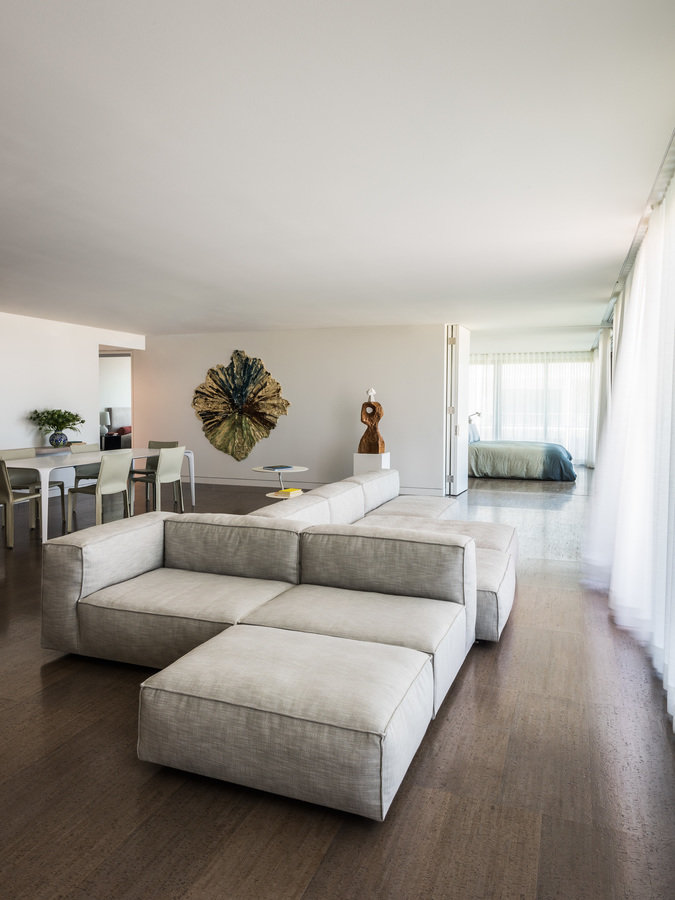 Open living room and dining room with view into bedroom. Dark cork floor, white walls, beige modern sofa and sheer curtains.