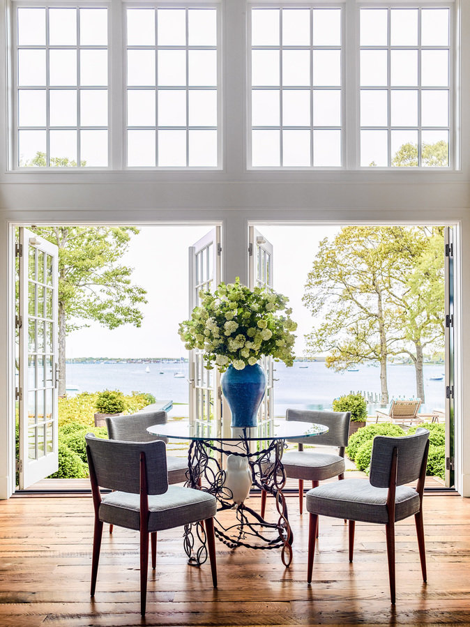 Glass-top café table with chairs, blue vase of white flowers, high divided light windows and lower French doors open to bay.