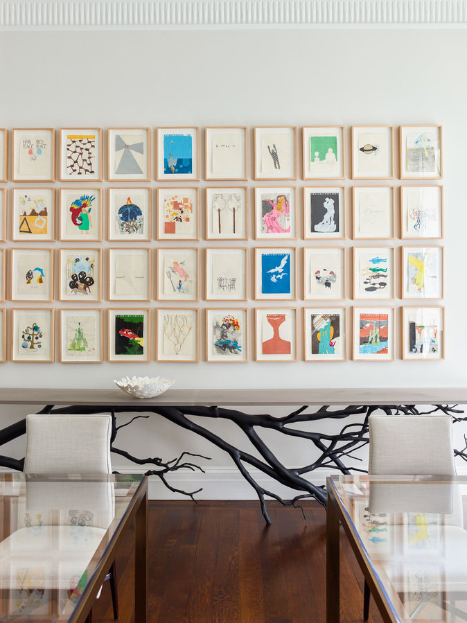Wall of framed art in a grid over a long table with marble top and metal tree branch base and two glass tables in front.