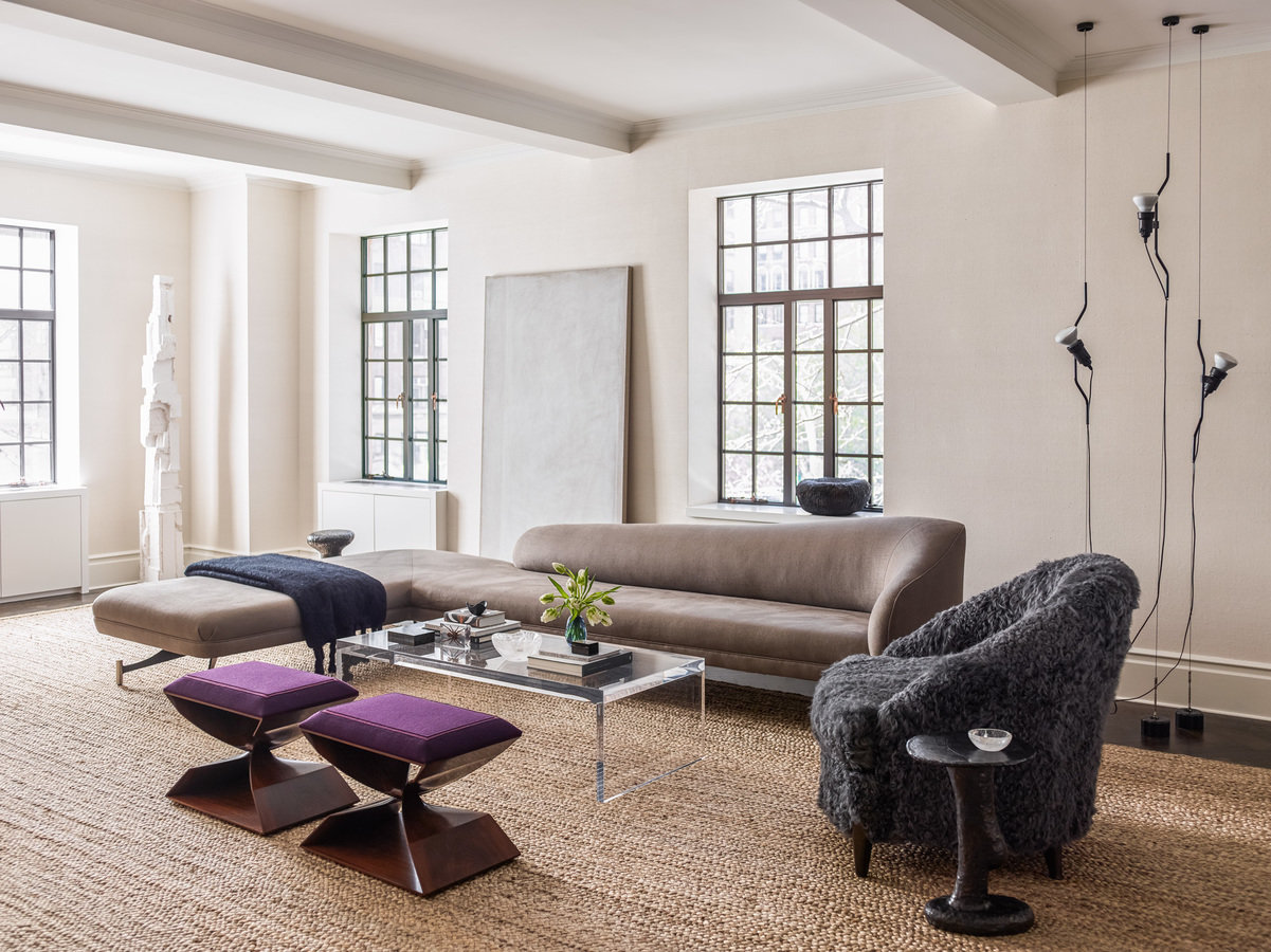 Beige-walled living room with sisal rug, casement windows, and a mix of modern and antique seating and a Lucite coffee table.