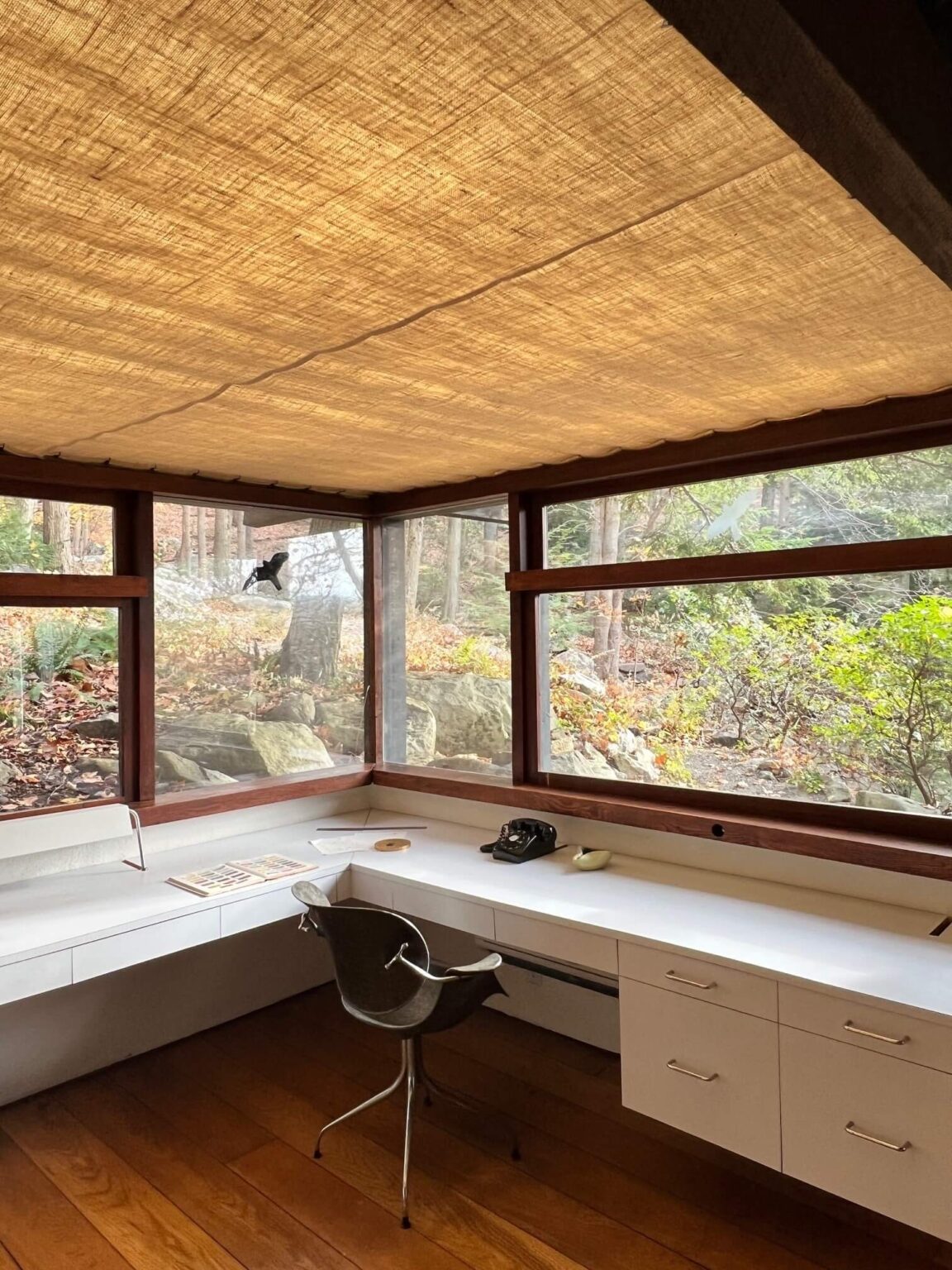 A home office space with window walls, a luminescent linen ceiling, a white wrap-around desk and a vintage Herman Miller chair.