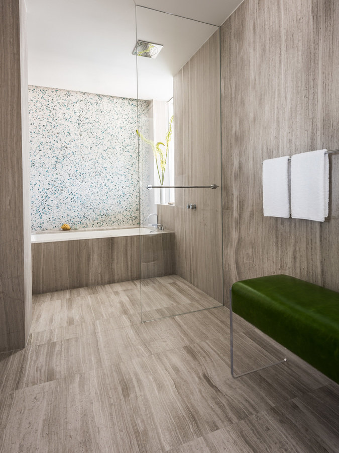 Modern bathroom with limestone walls, floor and tub; shower with glass door and terrazzo wall, lucite bench with green seat.