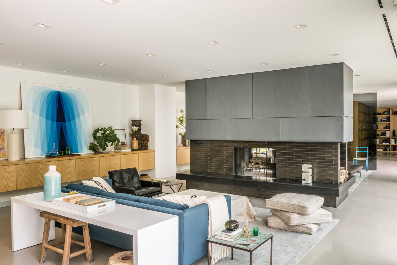 Great room divided by a two-sided fireplace in central wall, seating area with desk and stool and wall of low wood cabinets.