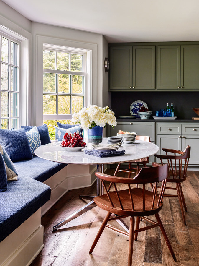 Window seat with blue cushion, white breakfast table and wood chairs in kitchen with green and white cabinets and wood floor.