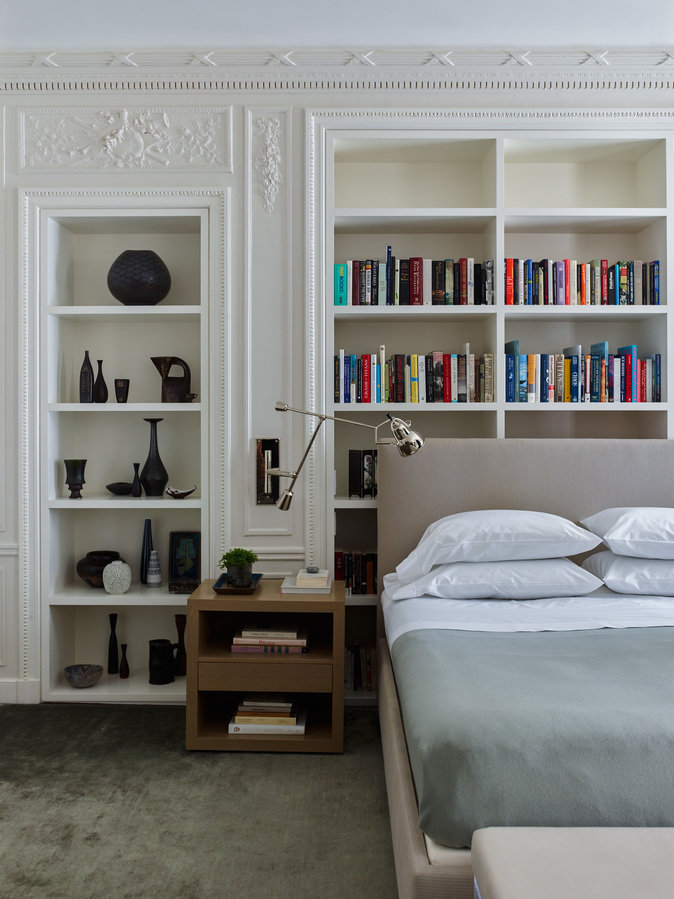 Bedroom with prewar details, shelf niches with black vases and books, upholstered bed, nightstand, and wall-mounted lamp.