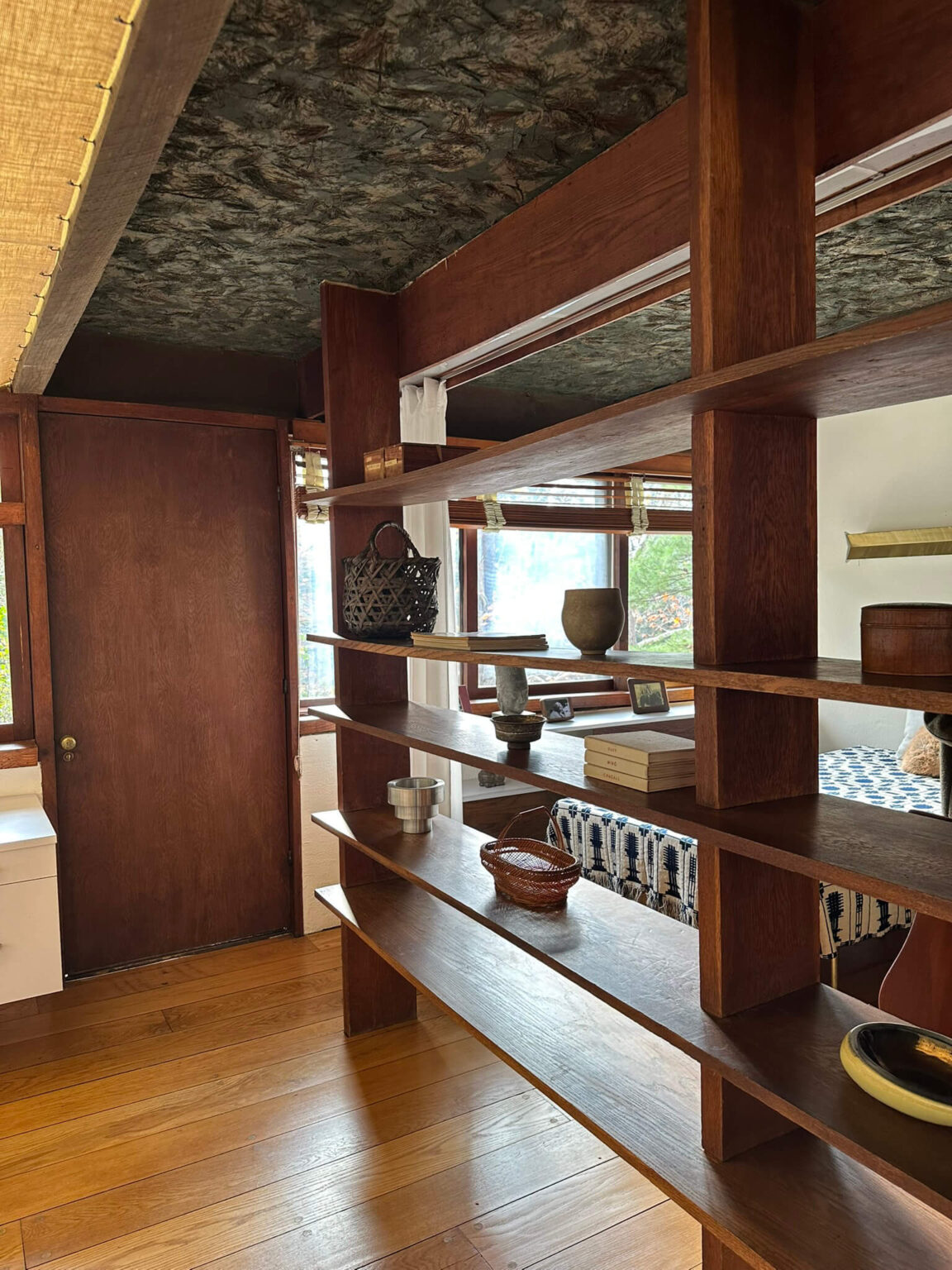 Interior with wood floor, decorative pine needle ceiling and an open wood shelf with a bedroom in the background.