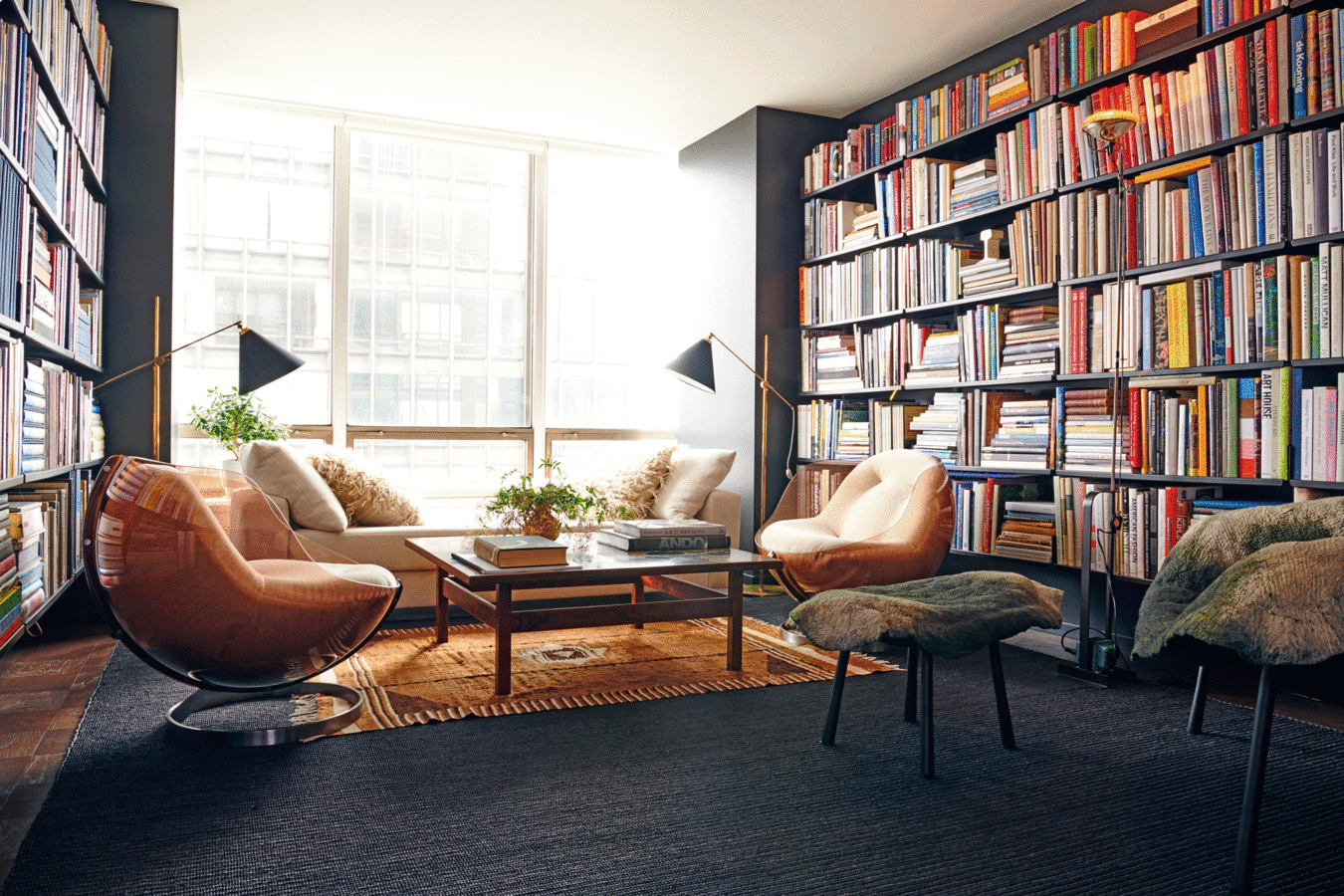 Library/guest room with window wall, two facing walls of floor-to-ceiling books, sofa, floor lamps and vintage armchairs.