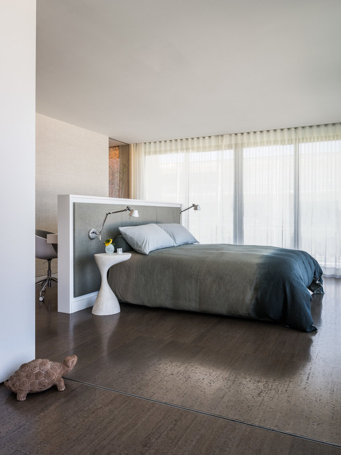 Bedroom with dark cork floors, wall of windows with sheer white shades, bed with partition wall headboard with sconces.