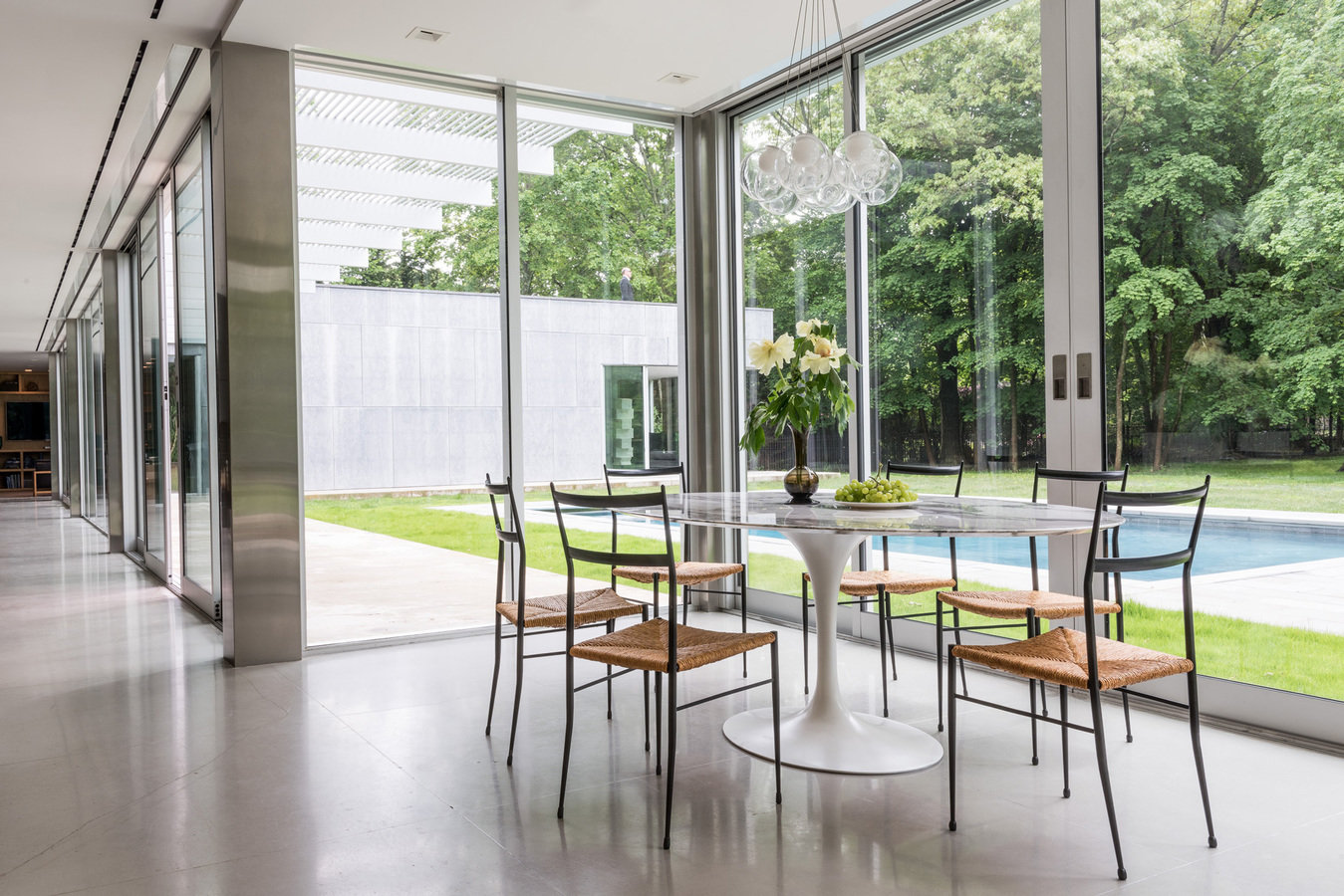 Glass-walled room with white Saarinen table with metal and rattan chairs, glass globe chandelier, view to pool in backyard.