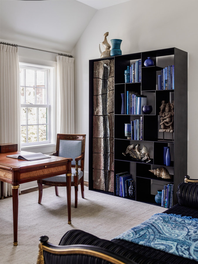Study with antique wood desk and chair near window with white curtains, blackened steel bookcase with books and sculptures.