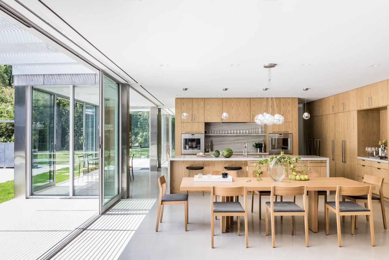 Glass-walled side of house with modern dining table and chairs and kitchen in background in light wood and stainless steel.