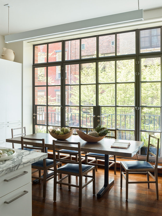 Rear wall of kitchen with casement doors, modern stone and metal dining table, wood chairs with upholstered leather seats.