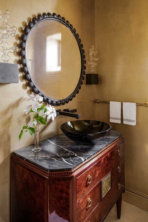 Powder room with gold walls and an antique dark wood dresser with black marble top and sink basin and round mirror.