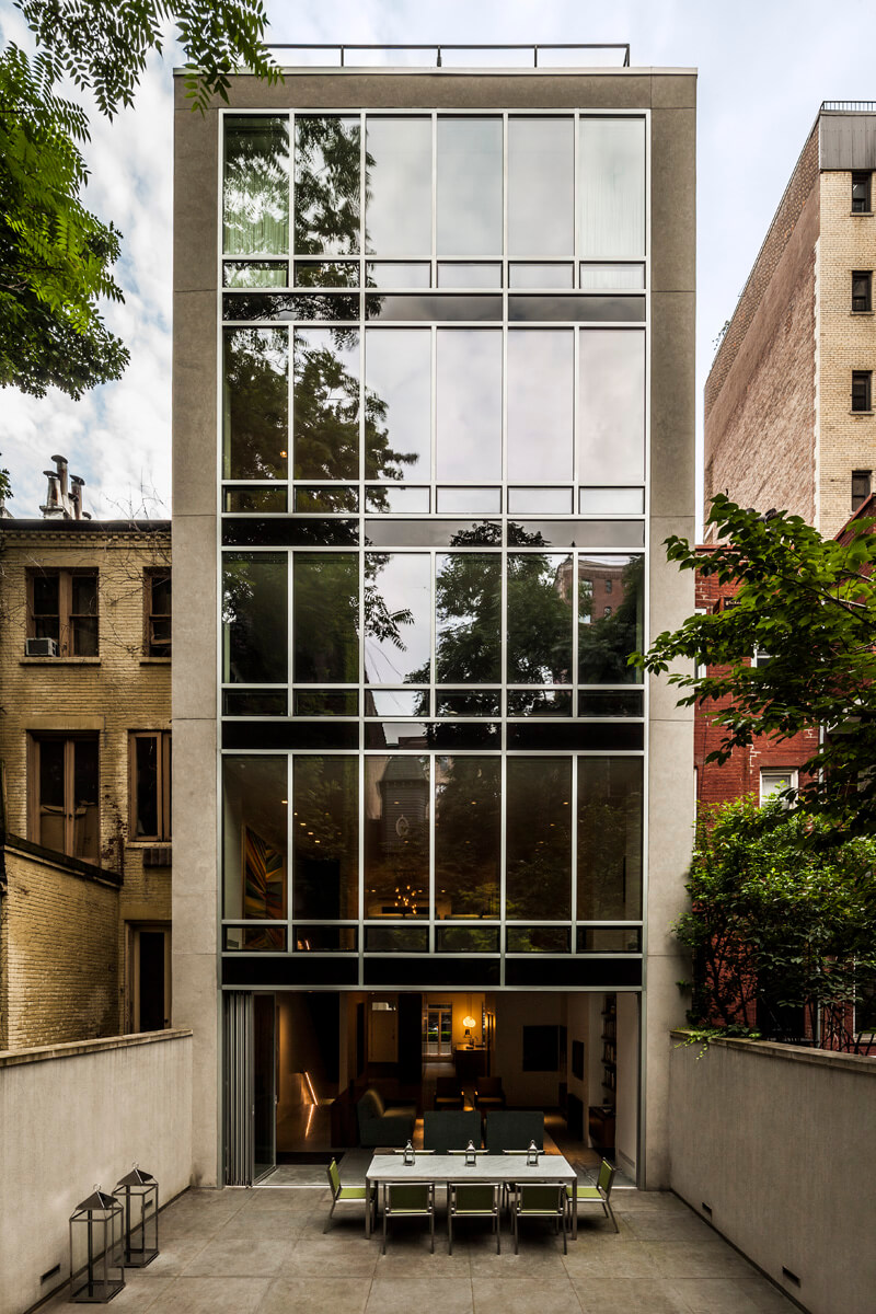 Rear facade of a townhouse with floor-to-ceiling windows. Open ground floor and a dining table on the back patio.