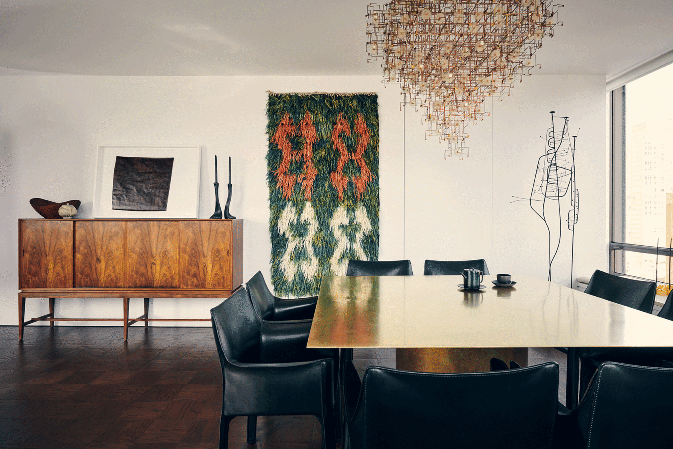 Dining room with brass table, black leather chairs, a bronze and dandelion chandelier; wood cabinet and green wall-hanging.