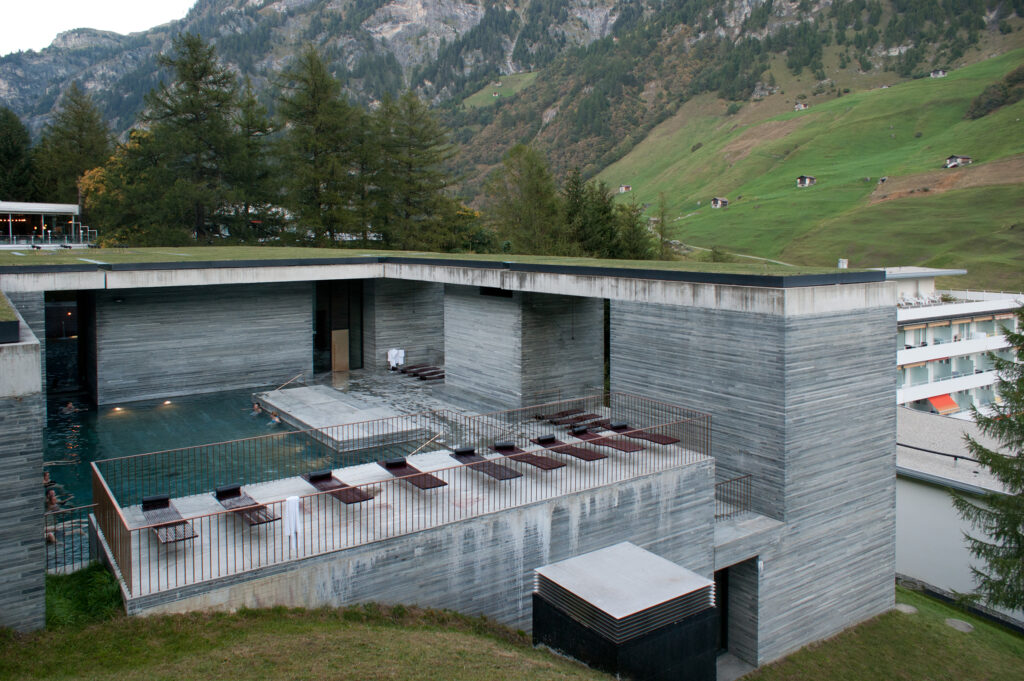 A modern stone building built into the landscape, with a pool and lounge chairs on the deck; mountains in the background.