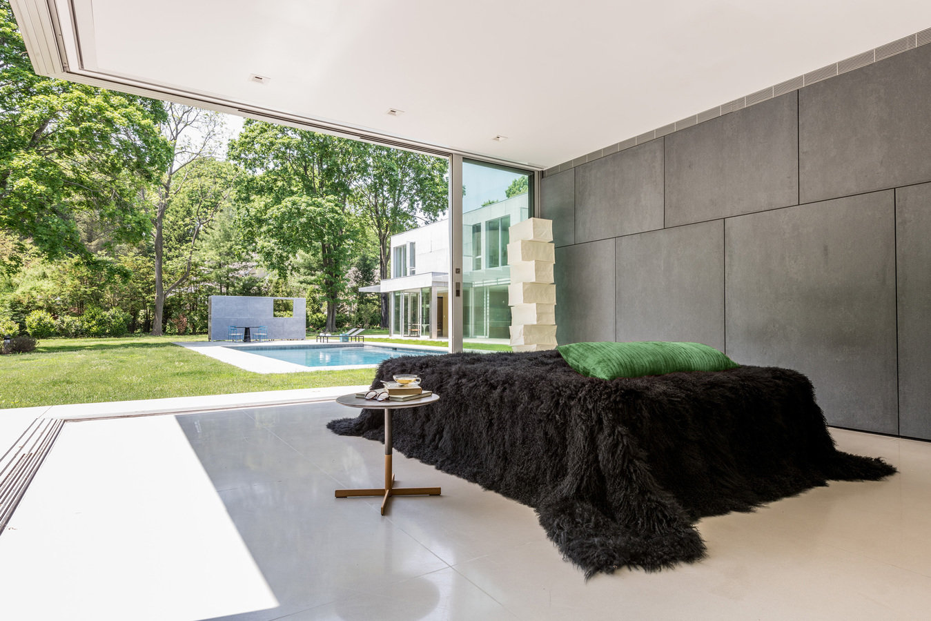 Primary bedroom with composite concrete panel wall, bed with black fur cover, sliding doors open to backyard with pool view.