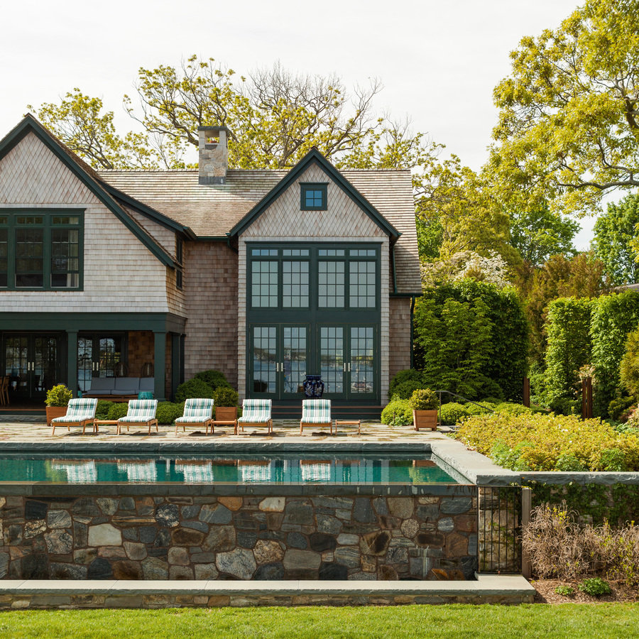 Back of wood-shingle house and patio with swimming pool and lounge chairs, surrounded by lush green landscaped yard.
