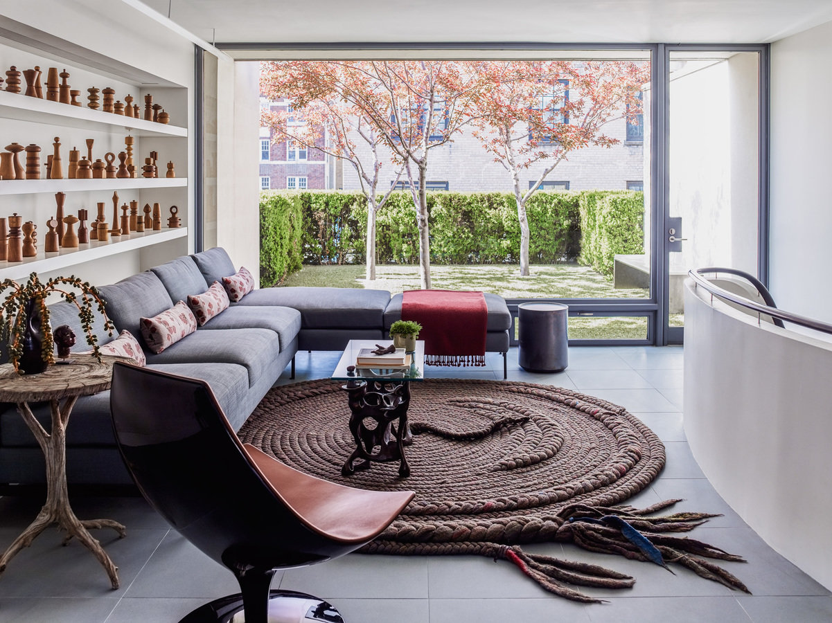 Living room with modern gray sofa, round rug, lounge chair, white shelves with figurines, full-wall window with garden view.