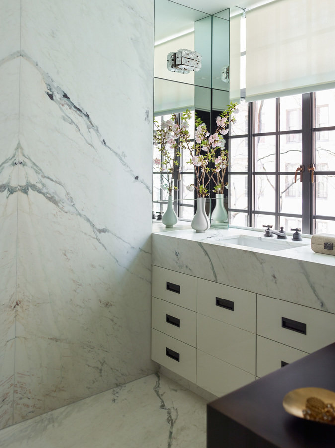 Corner of a white marble bathroom with sink vanity and white cabinetry, casement windows with white shade and mirror wall.