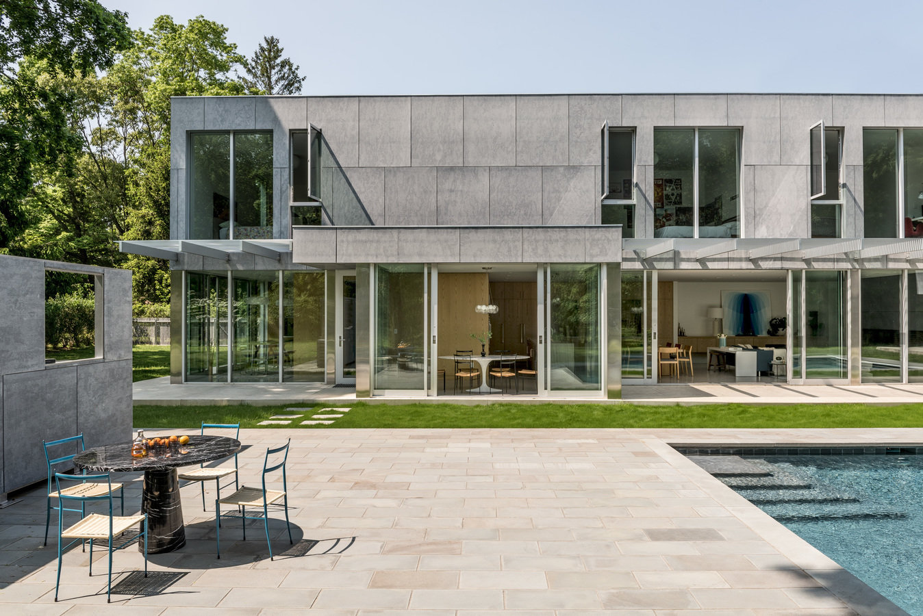 Rear façade of modern two-level house in cement and glass, surrounding lawn, stone patio with table and chairs, and pool.