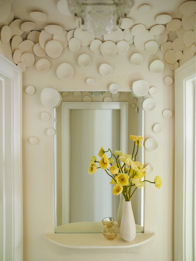 Entry foyer with off-white walls encrusted with plaster discs and a shelf with mirror holding a vase of yellow flowers.