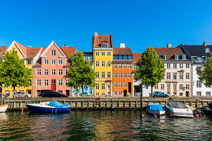 A row of brightly colored townhouses against a bright blue sky with a canal and boats in the foreground.