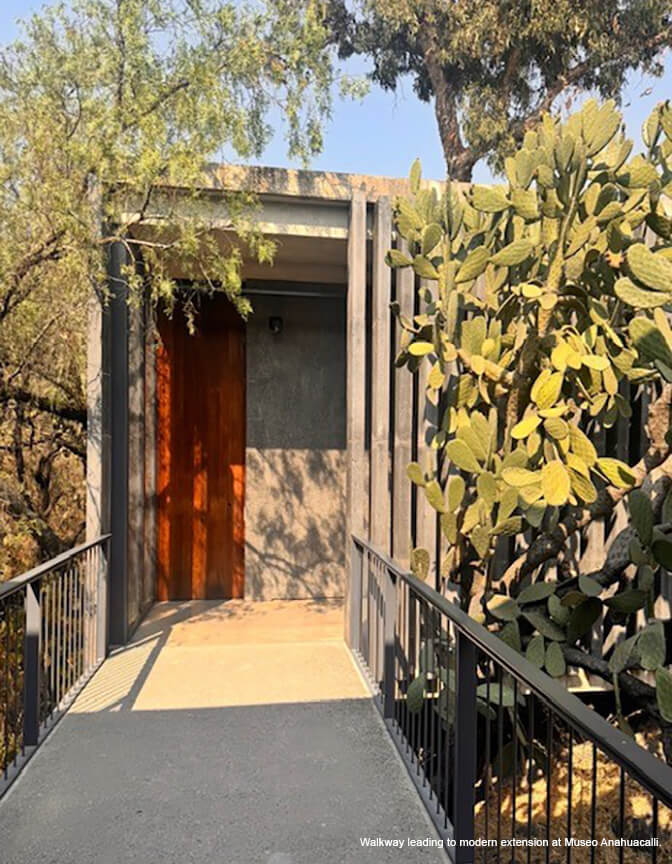 A concrete walkway with a railing leading to a building with a deep orange and concrete wall and wood slats; a cactus.
