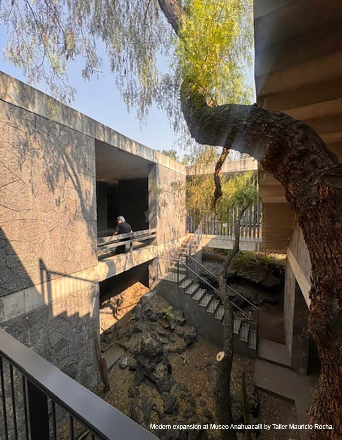 An outdoor courtyard surrounded by stone and concrete buildings with a staircase in the rear and a tree in the foreground.