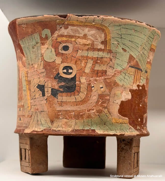 A short rounded sculptural vessel on three legs made of orange stone with painting in orange, beige, light green and black.