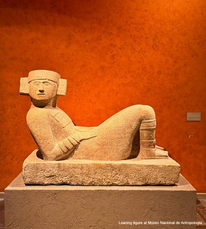 A large-scale sculpture made of beige stone of a person with a headpiece on, reclining. An orange wall in the background.
