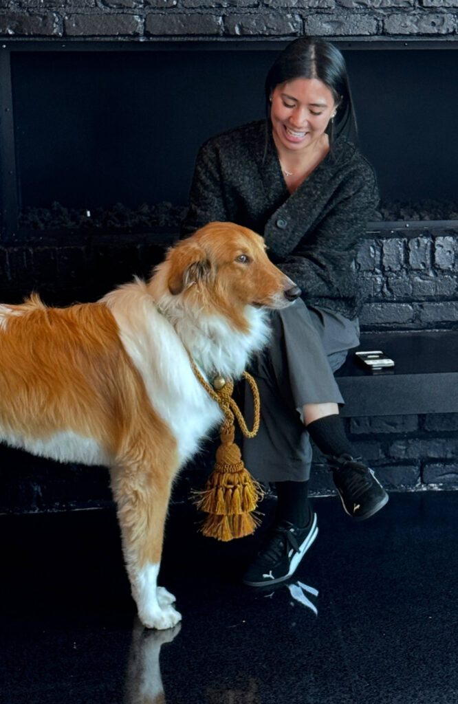 A woman with dark hair in dark gray clothing sitting in front of a black brick wall with a white and golden brown dog.