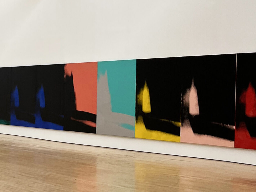 A gallery with a series of canvases side-by-side on a white wall with different versions of a similar painting in black and bright colors.