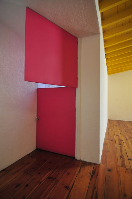A room with wood floors and white walls, a yellow ceiling to the right and a split plywood door in bright pink.