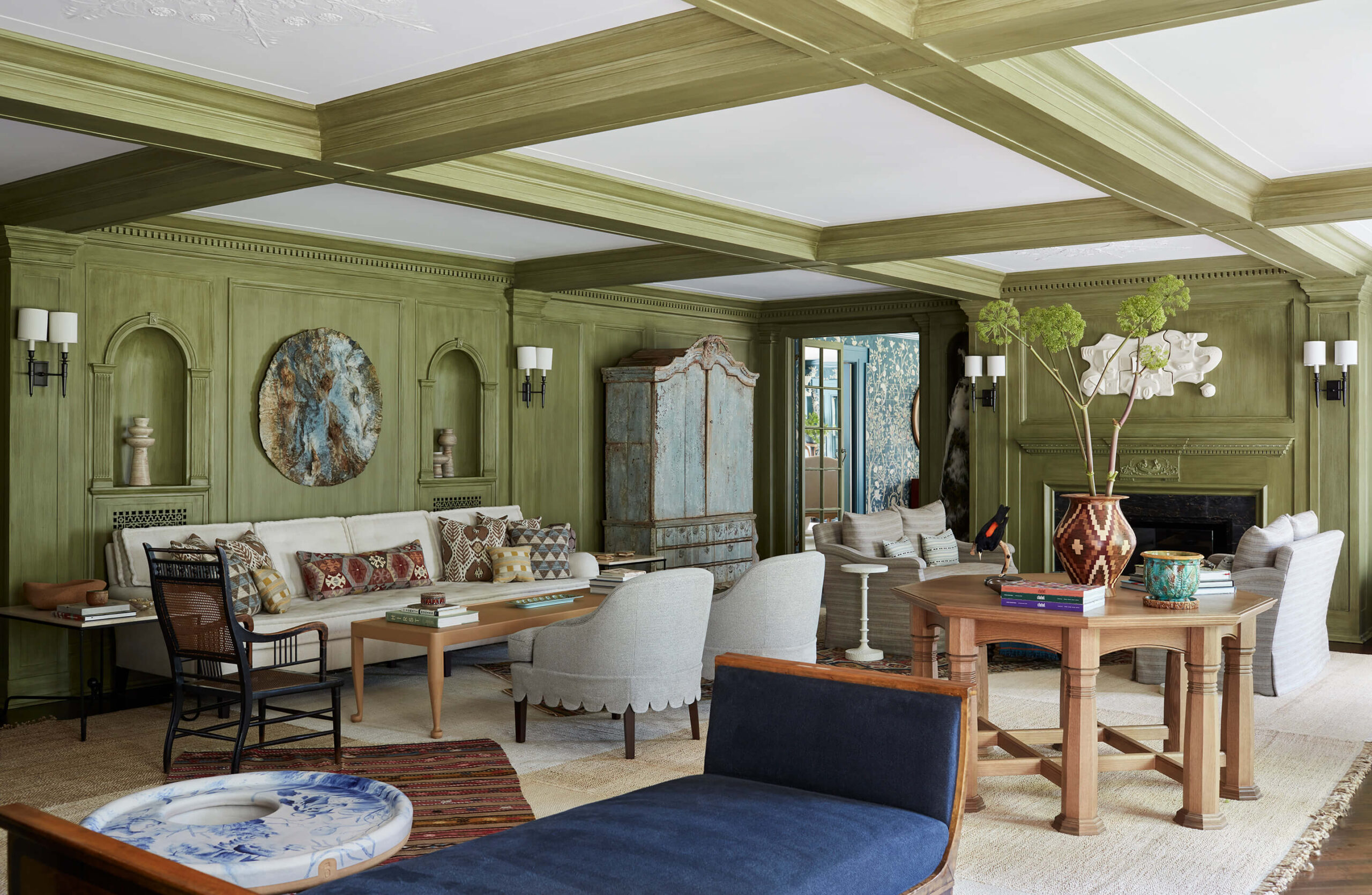 Large living room with green millwork walls, coffered ceiling, blue upholstered chaise, a sofa, and various chairs and tables.