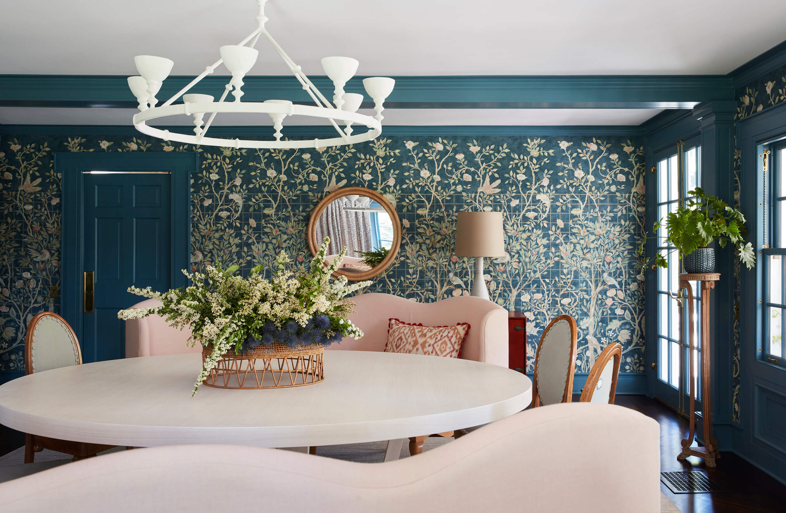 Dining room with round white table, white chandelier, pink upholstered bench seating and blue and white floral wallpaper.