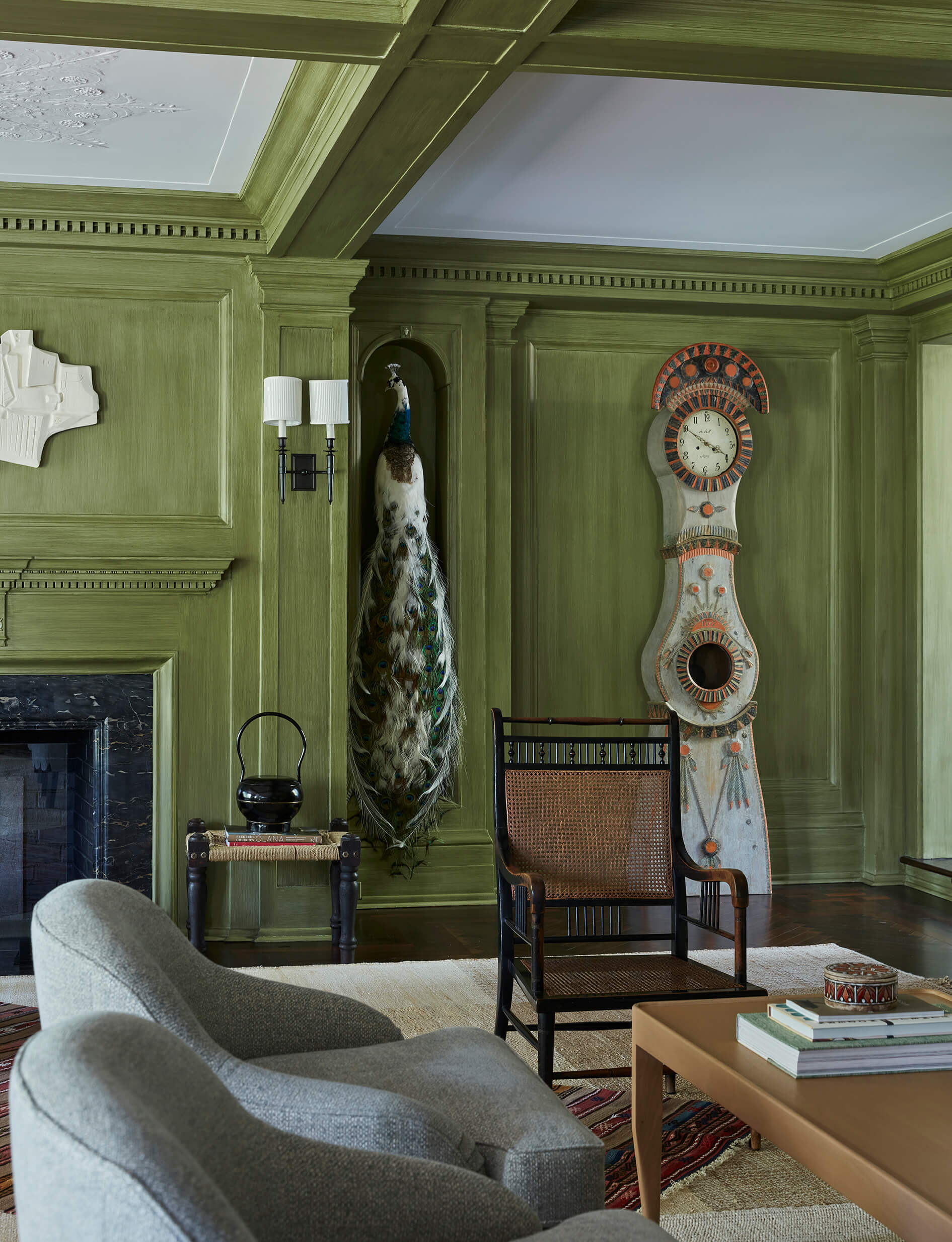 Room with green-painted millwork walls, standing clock and taxidermy peacock, cane chair, gray lounge chairs and coffee table.