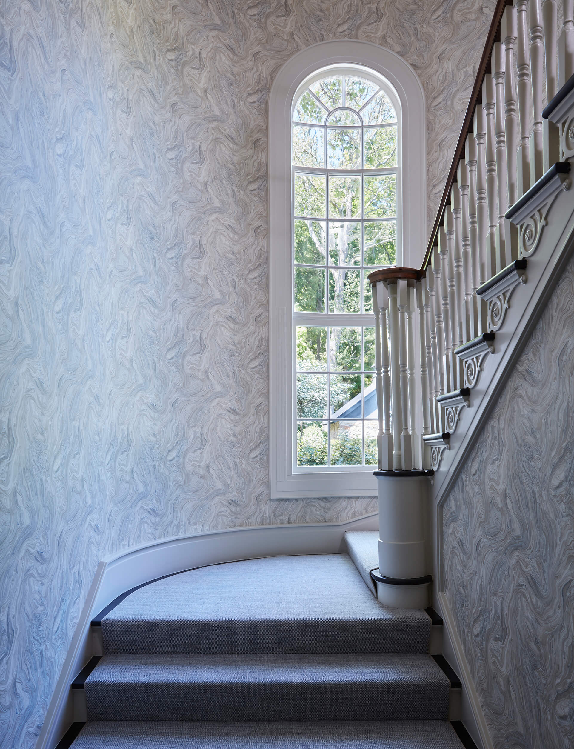 Landing of curved staircase with gray-blue carpet, blue-gray and white swirled wallpaper and narrow, white trimmed window.