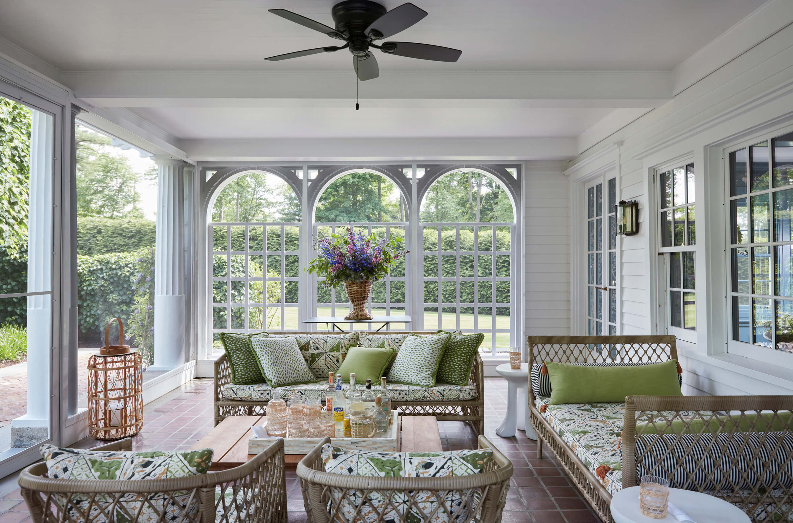 Enclosed porch with white siding, arched windows, white columns, ceiling fan and rattan furniture with white and green cushions.